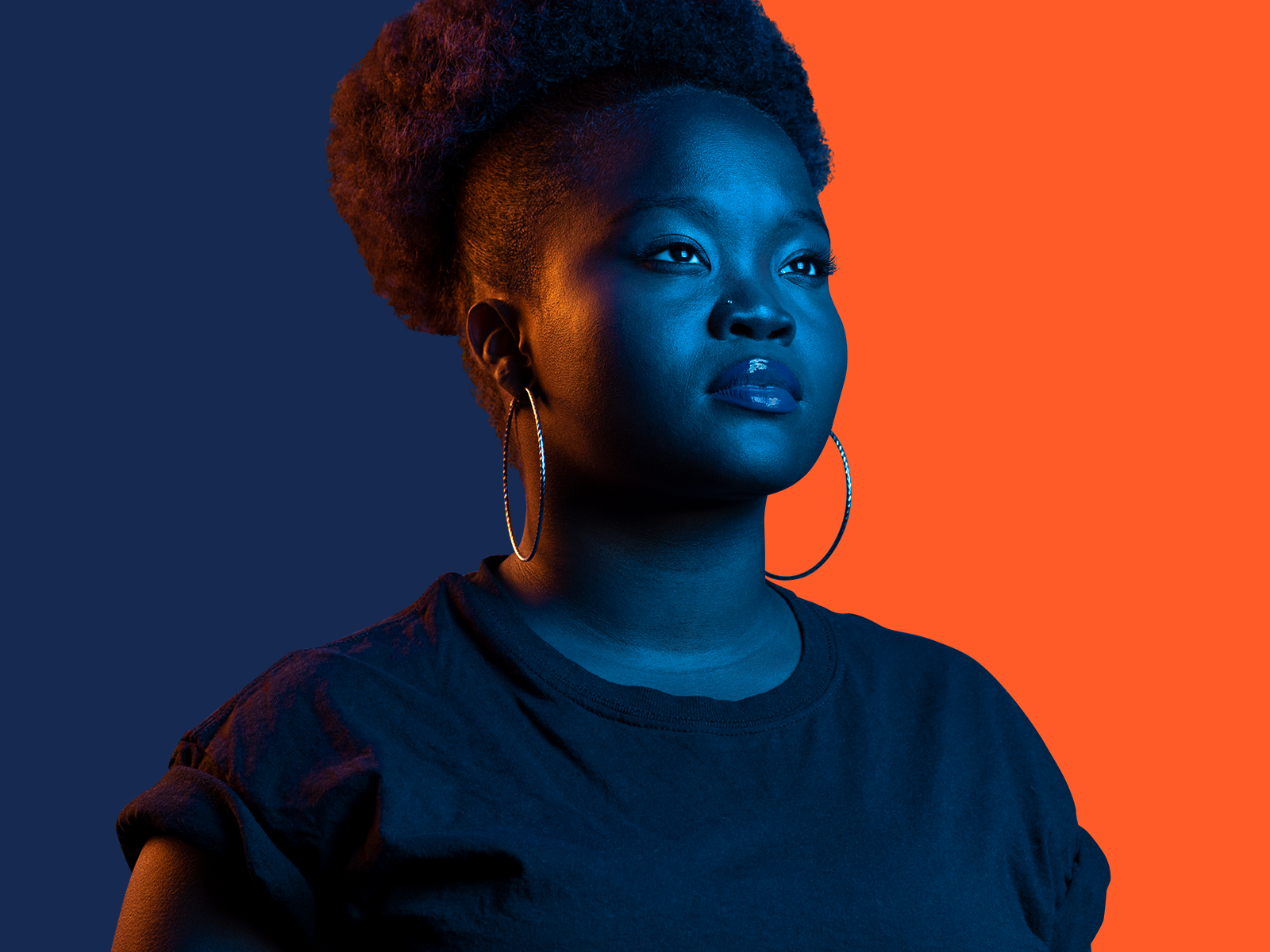 A Black woman stands against a blue and orange backdrop, with orange light against the blue side and blue light against the orange side.